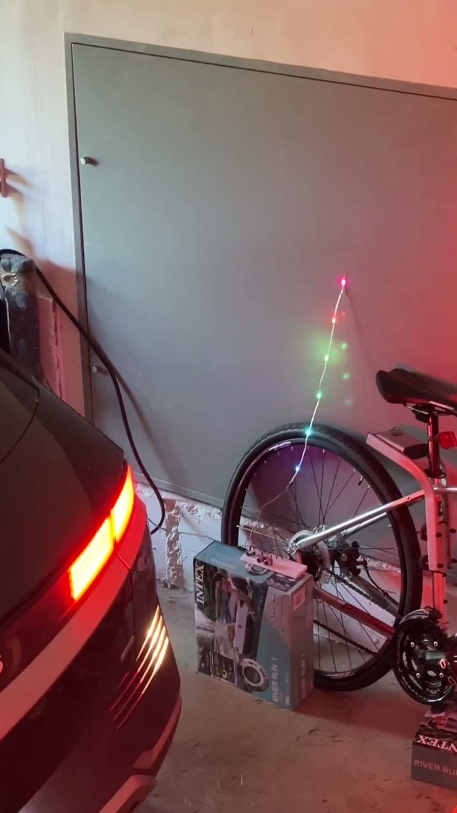 Inside a garage, a car backs slowly toward the wall, its tail lights washing the wall pink. A bicycle leans against the wall beside a cardboard box with a small circuit board on it, and a thin string of LEDs running up the wall lights up in green, blue and red as the car gets close.