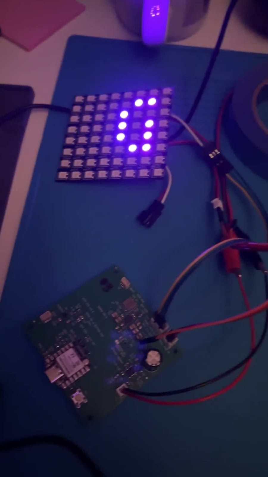 A green circuit board lies on a blue silicone work mat, wired with red and black clip leads to a separate square grid of RGB LEDs. The grid cycles through letters and patterns in blue, yellow, red and green while a gloved hand reaches in at the bottom of the frame.