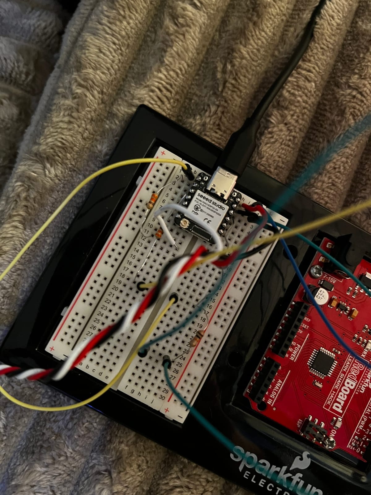 A breadboard on a black plastic mount resting on a couch. A red Arduino Uno (SparkFun RedBoard) at the top is wired by blue, yellow, red and black jumper wires down to a white breadboard holding a small black Seeed Studio XIAO ESP32, a USB-C cable, and a few loose resistors. An in-progress test of the lantern's LED controller.