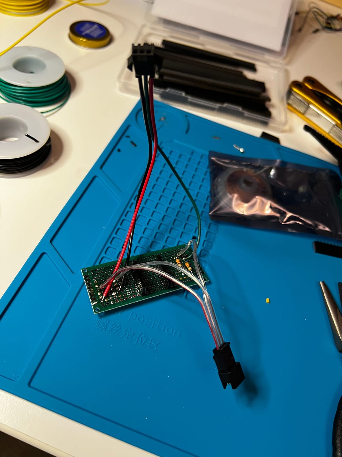 A small hand-soldered green protoboard sits on a blue silicone soldering mat. A black 3-pin connector with red, white, and black wires is soldered to one end, feeding an addressable LED strip, with a separate red-and-black power lead. Around it are spools of green and yellow hookup wire, flush cutters, and pliers.