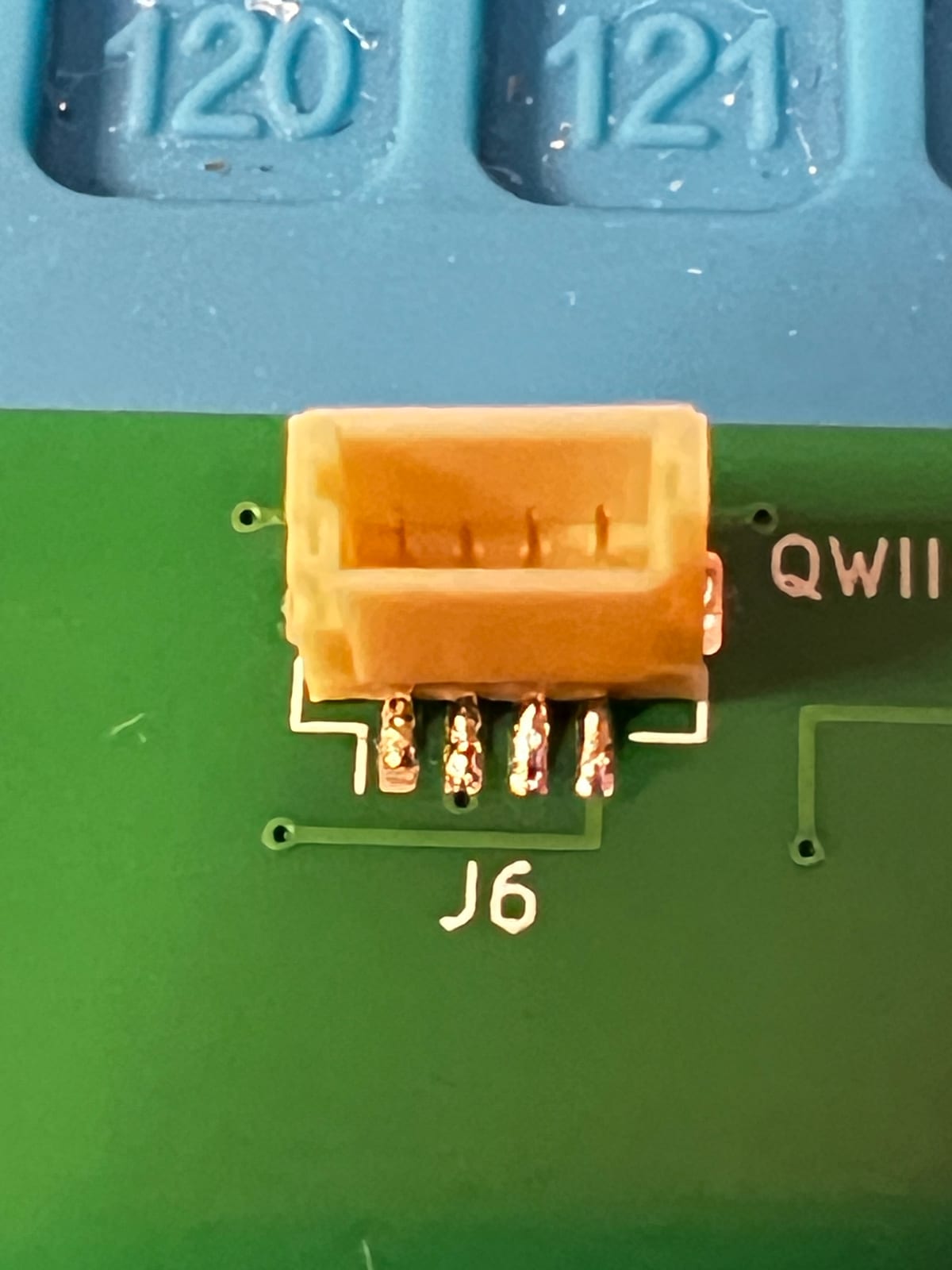 Extreme close-up of a small cream-coloured four-pin Qwiic connector soldered onto a green circuit board, its four solder joints bright against the copper pads, with the silkscreen label J6 beside it and a blue silicone work mat behind.