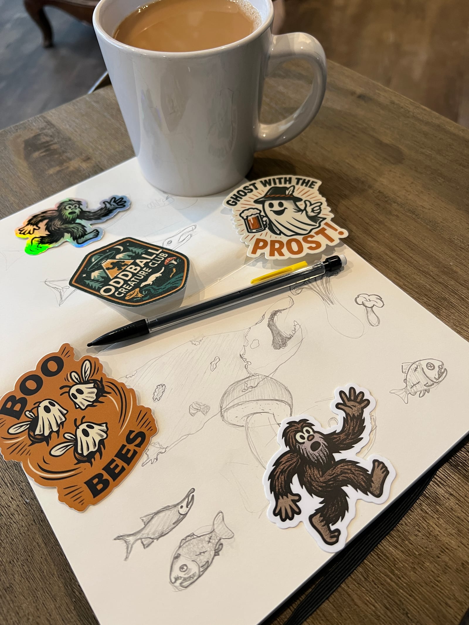 Oddball Creature Club stickers and an embroidered patch on a sketchbook of pencilled creatures, beside a coffee mug